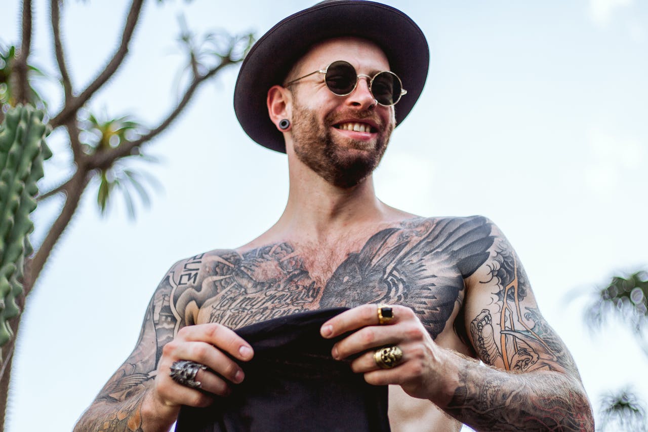 A fashionable tattooed man poses confidently outdoors, wearing a hat and sunglasses. Perfect for lifestyle and fashion themes.