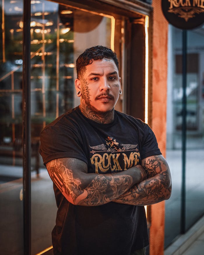 Portrait of a tattooed man in rock-themed fashion standing confidently with crossed arms in an urban setting.